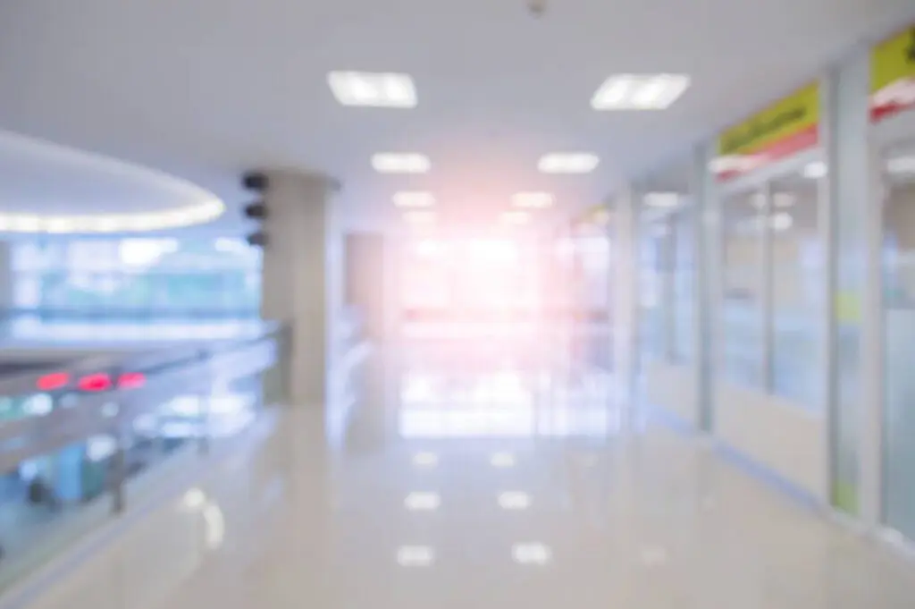 Blurred image of a bright, empty hallway with reflective floors, large windows, and overhead lights, possibly in a hospital or modern office building.